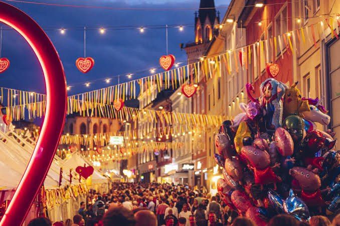 Eine überfüllte Straße mit bunten Luftballons und Herzdekorationen, unter einem Nachthimmel. Gebäude säumen die Straße, geschmückt mit Lichtern und Flaggen.