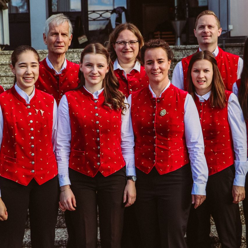 Eine Gruppe von Menschen in roten Westen mit silbernen Knöpfen steht auf den Stufen eines Gebäudes. Sie lächeln und posieren für ein Foto. Die Frau auf der rechten Seite trägt eine Brille. Hinter ihnen gibt es eine verschwommene Ansicht eines Gebäudes und eines Zauns.