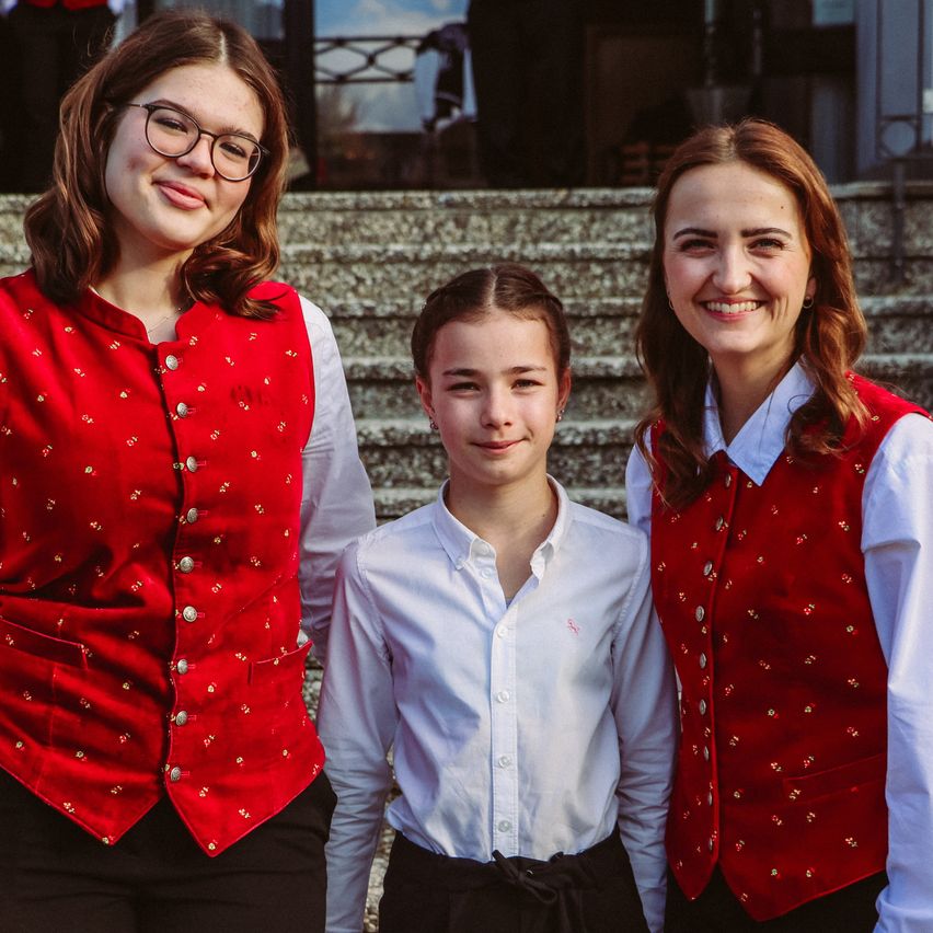 Drei junge Frauen posieren auf einer Treppe für ein Foto, alle tragen rote Westen. Das Mädchen in der Mitte trägt ein weißes Hemd.