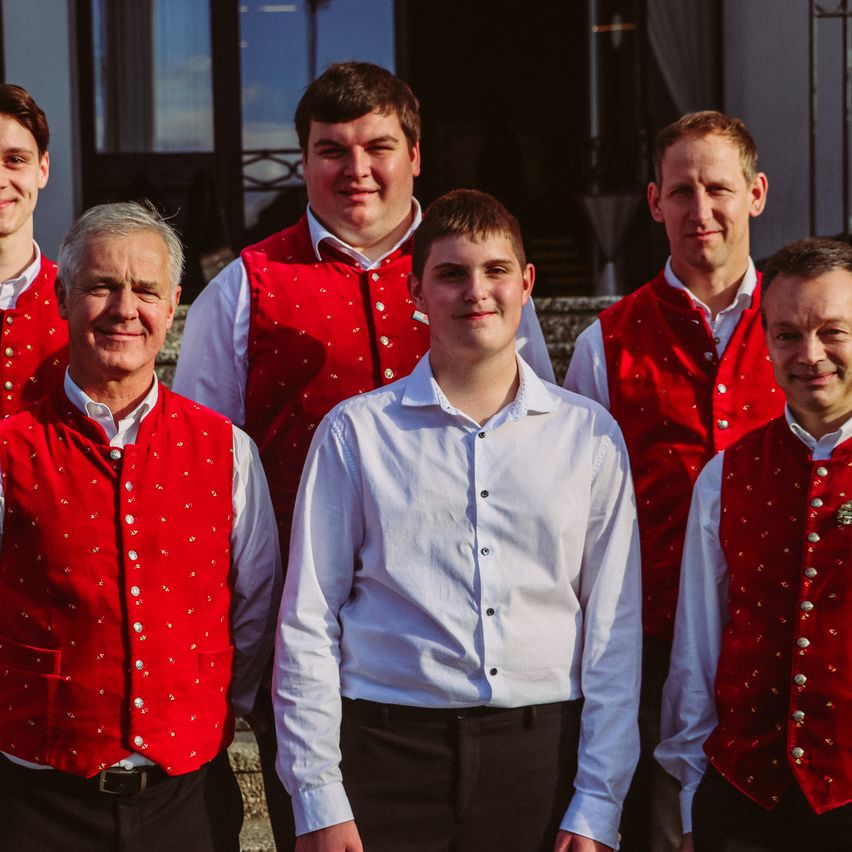 Sieben Männer in roten Westen und weißen Hemden stehen nebeneinander und lächeln für die Kamera.