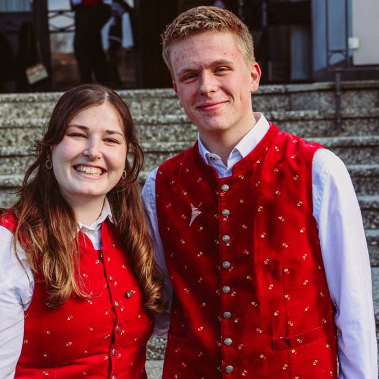 Ein junger Mann und eine junge Frau in roten Westen lächeln in die Kamera, während sie vor einem Gebäude auf einer Treppe stehen.