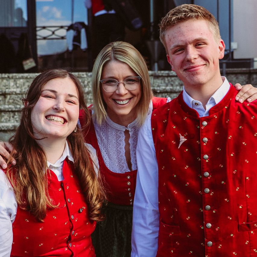 Drei Menschen stehen zusammen auf einer Treppe. Zwei Frauen und ein Mann lächeln für die Kamera. Die beiden Frauen tragen rote Westen. Der Mann trägt eine rote Weste mit einem weißen Hemd.