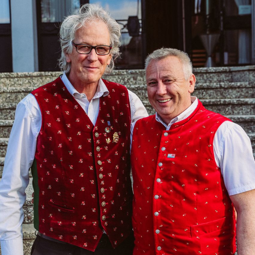 Zwei Männer stehen auf einer Treppe, einer mit einer Brille und einem weißen Hemd, der andere mit einem roten Weste mit Knöpfen und einem weißen Hemd, beide lächeln für die Kamera.