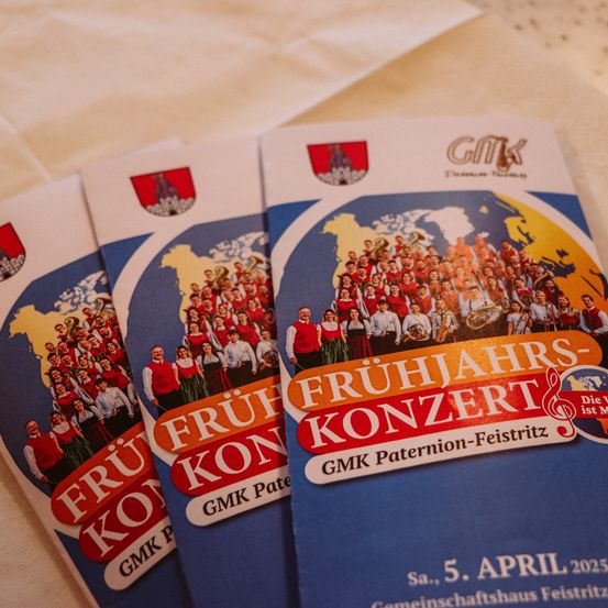 Drei Broschüren für ein Frühlingskonzert am 5. April 2025 mit einem Orchester, einer Gruppe von Musikern und einer Weltkarte im Hintergrund. Die Broschüre enthält das GMK Paternion-Feistritz-Logo und eine blau-weiße Titelseite.