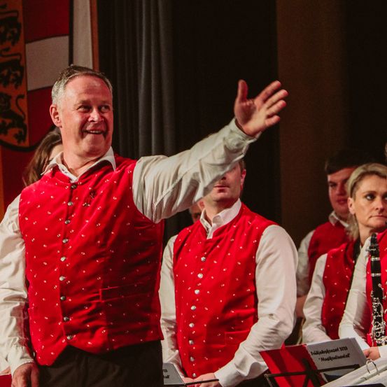 Eine Gruppe von Musikern auf der Bühne, in roten Westen, führt ein Musikkonzert auf. Der Mann vorne winkt mit der Hand.