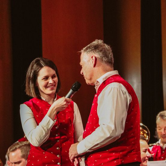 Eine Frau und ein Mann in roten Westen stehen auf der Bühne mit einem Mikrofon. Sie scheinen bei einer Veranstaltung aufzutreten.