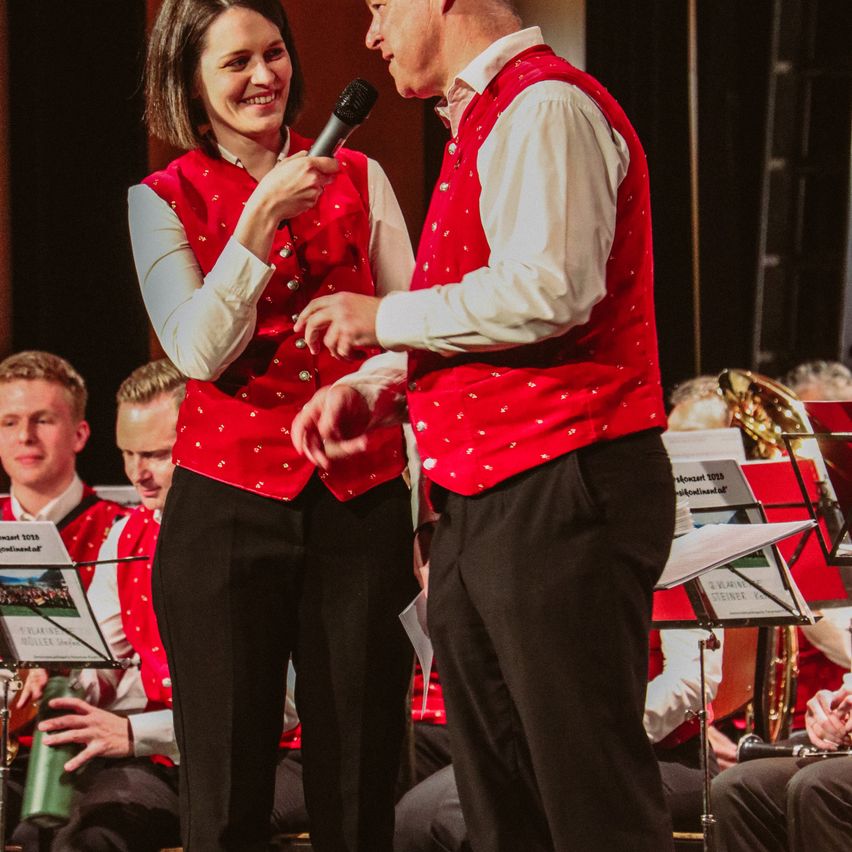 Eine lächelnde Frau in einem roten Westchen hält ein Mikrofon, während ein Mann in einer ähnlichen Weste neben ihr steht, möglicherweise bei einer musikalischen Veranstaltung. Andere Musiker spielen hinter ihnen.