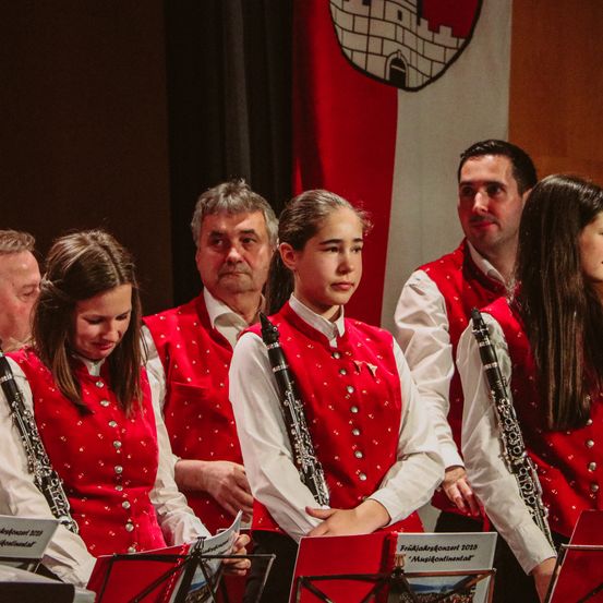 Eine Gruppe von Musikern in roten Westen mit silbernen Knöpfen, die Klarinetten halten und etwas vor sich betrachten.