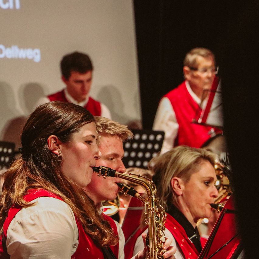 Eine Gruppe von Musikern, darunter zwei Frauen, die Saxophon spielen, tritt auf der Bühne auf. Der Mann hinter ihnen hält eine Trompete. Sie tragen alle rote Westen und weiße Hemden. Im Hintergrund befindet sich ein Bildschirm mit dem Wort Dellweg. Das Publikum ist verschwommen.
