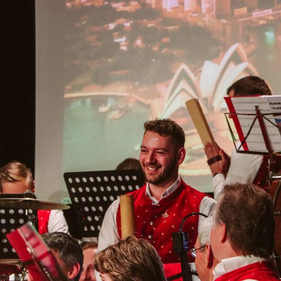 Eine Gruppe von Musikern in roten Westen, die vor einer großen Leinwand mit dem Opernhaus von Sydney auftreten.