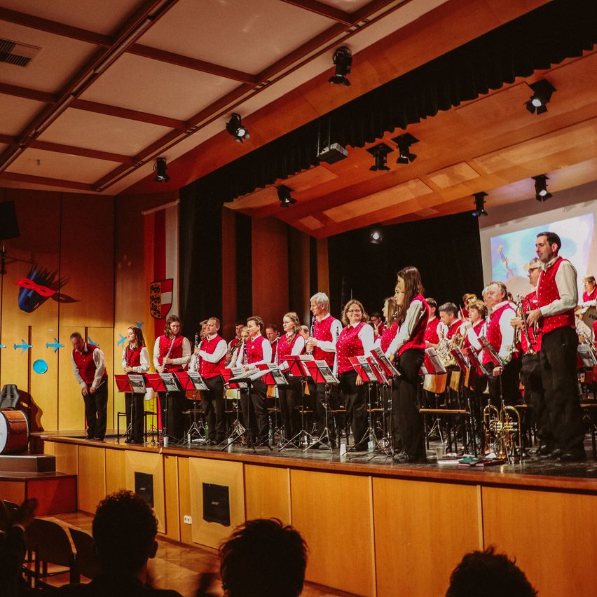 Ein Blechbläserensemble auf der Bühne mit roten Westen, Musikinstrumenten und einer Trommel. Eine Projektionsleinwand befindet sich hinter ihnen an der Wand.