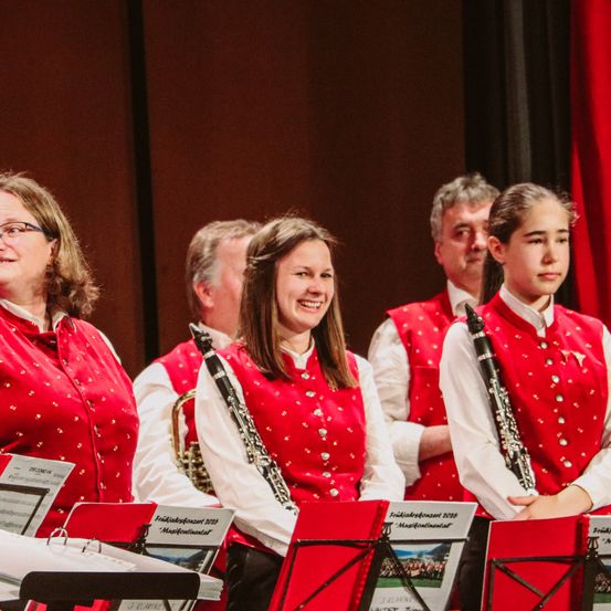 Eine Gruppe von Musikern auf der Bühne bei einer musikalischen Veranstaltung. Drei Frauen und zwei Männer in roten Westen mit Silberpunkten. Sie spielen Musikinstrumente, darunter eine Trompete und eine Klarinette.