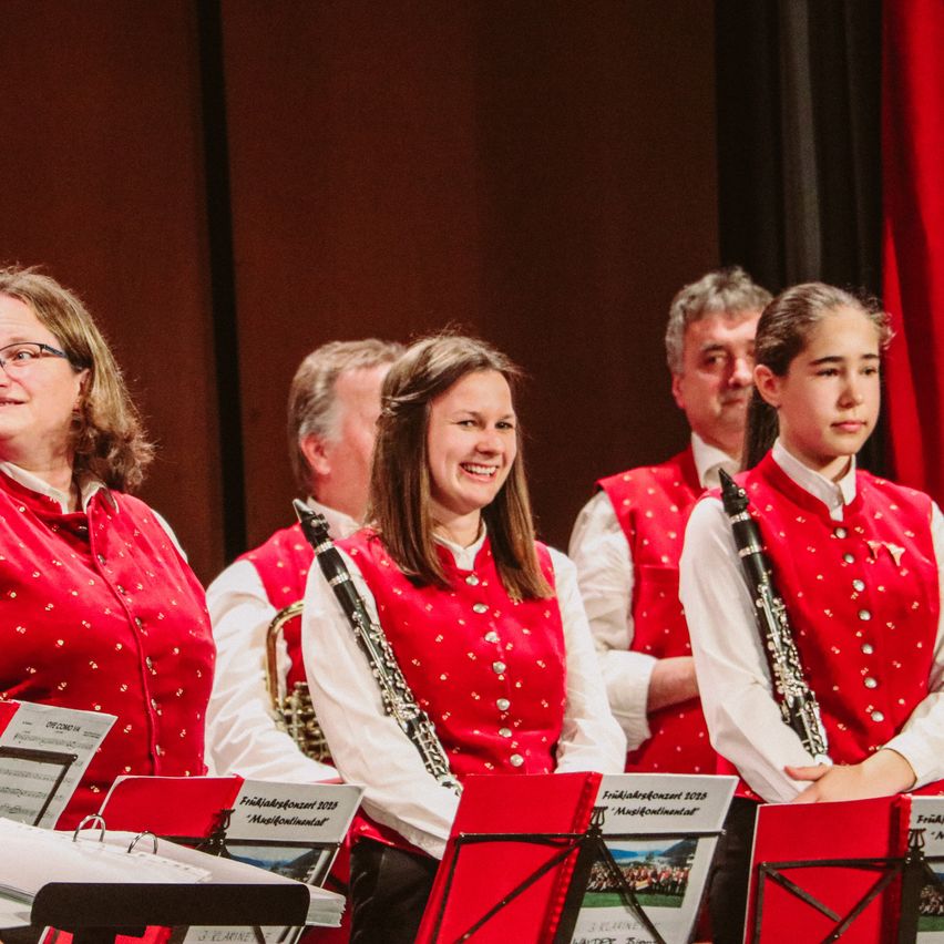 Eine Gruppe von Musikern auf der Bühne bei einer musikalischen Veranstaltung. Drei Frauen und zwei Männer in roten Westen mit Silberpunkten. Sie spielen Musikinstrumente, darunter eine Trompete und eine Klarinette.