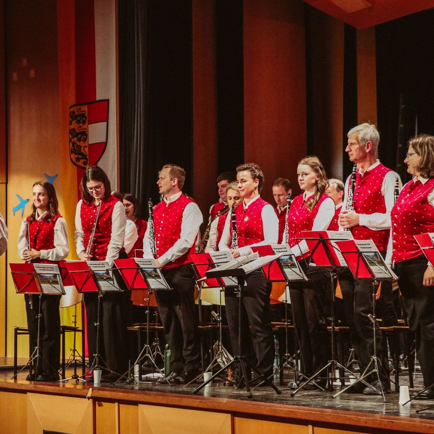 Eine Gruppe von Musikern steht auf der Bühne und hält ihre Instrumente, mit roten Notenständern vor ihnen und einer Person auf der linken Seite. Hinter ihnen befindet sich ein Bühnenvorhang und ein Logo an der Wand.
