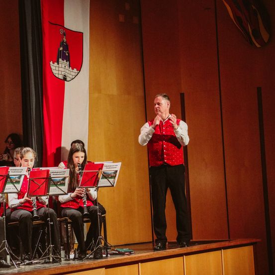 Eine Band von Kindern in roten Westen spielt ihre Instrumente mit einem Mann vor ihnen auf einer Bühne mit einer Fahne dahinter.