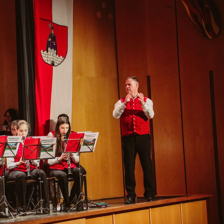 Eine Band von Kindern in roten Westen spielt ihre Instrumente mit einem Mann vor ihnen auf einer Bühne mit einer Fahne dahinter.