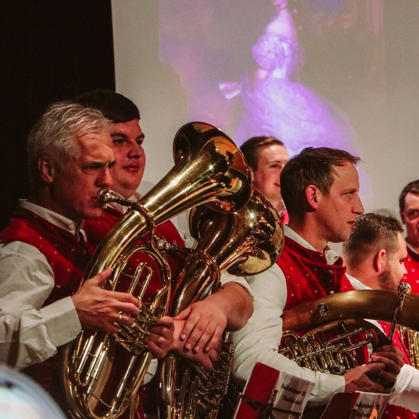 Eine Gruppe von Musikern spielt Blechblasinstrumente auf der Bühne. Sie tragen rote Westen mit weißen Hemden. Hinter ihnen befindet sich eine Projektion einer Frau in einem weißen Kleid auf einer weißen Leinwand.