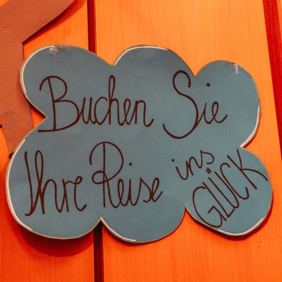 Ein blaues, wolkenförmiges Schild auf einer orangefarbenen Oberfläche mit der Aufschrift 'Buchen Sie Ihre Reise mit Glück' in deutscher Sprache.