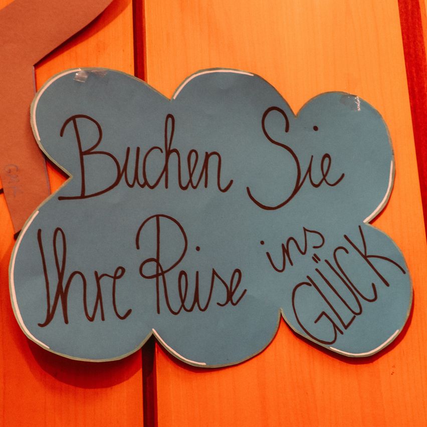 Ein blaues, wolkenförmiges Schild auf einer orangefarbenen Oberfläche mit der Aufschrift 'Buchen Sie Ihre Reise mit Glück' in deutscher Sprache.