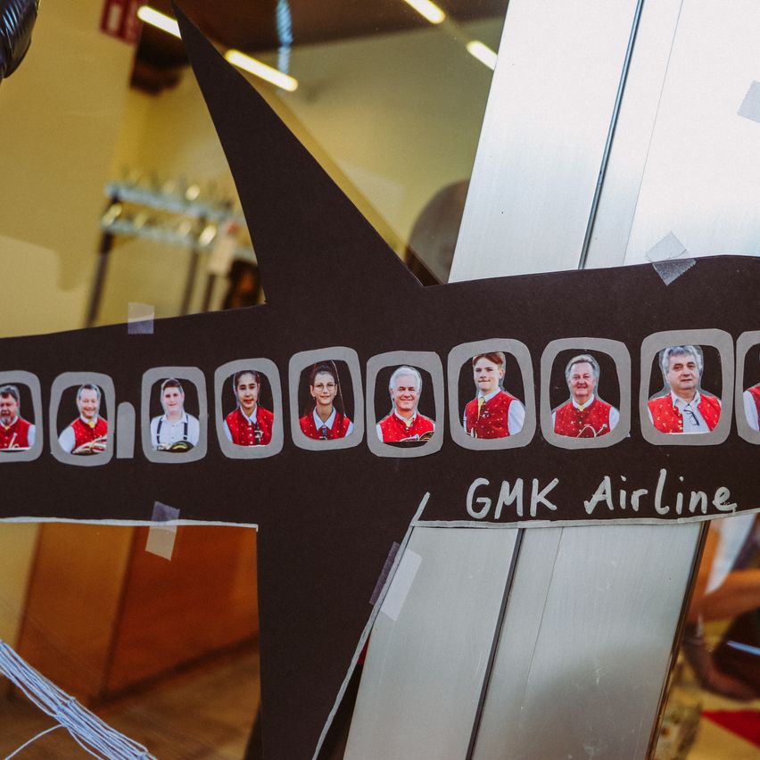 Eine Ausschnittform eines Flugzeugs mit neun Gesichtern von Erwachsenen in roten Westen ist mit GMK Airline beschriftet.