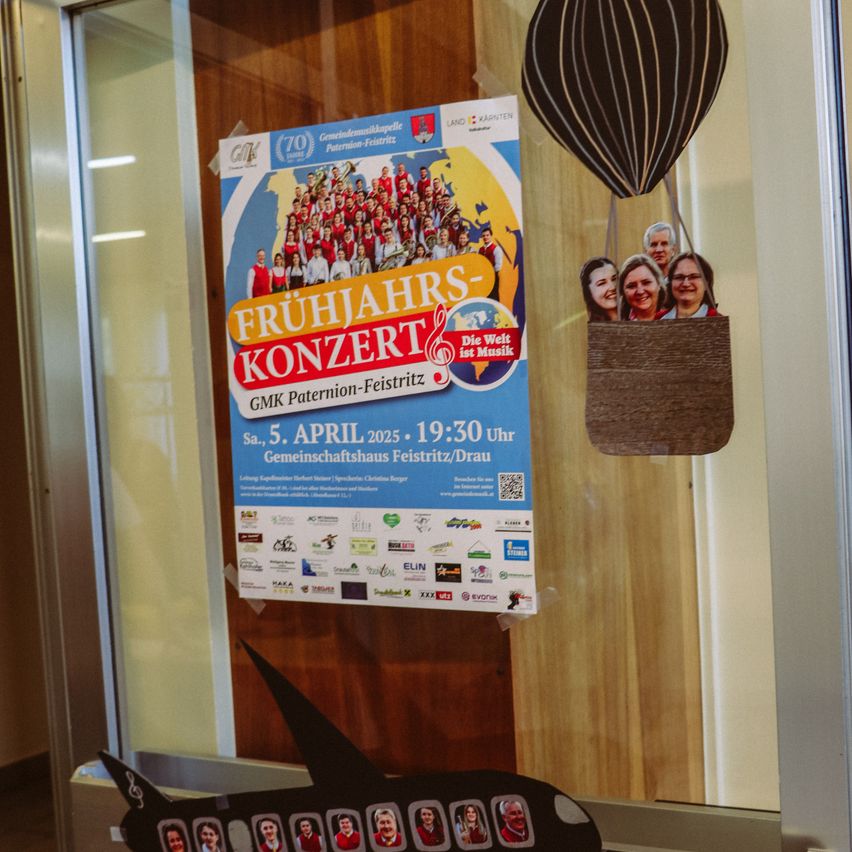 Plakat an einer Glastür mit einem Heißluftballon-Papierausschnitt. Das Plakat bewirbt ein Frühlingskonzert am 5. April 2025 um 19:30 Uhr im Gemeindehaus Feistritz/Drau.