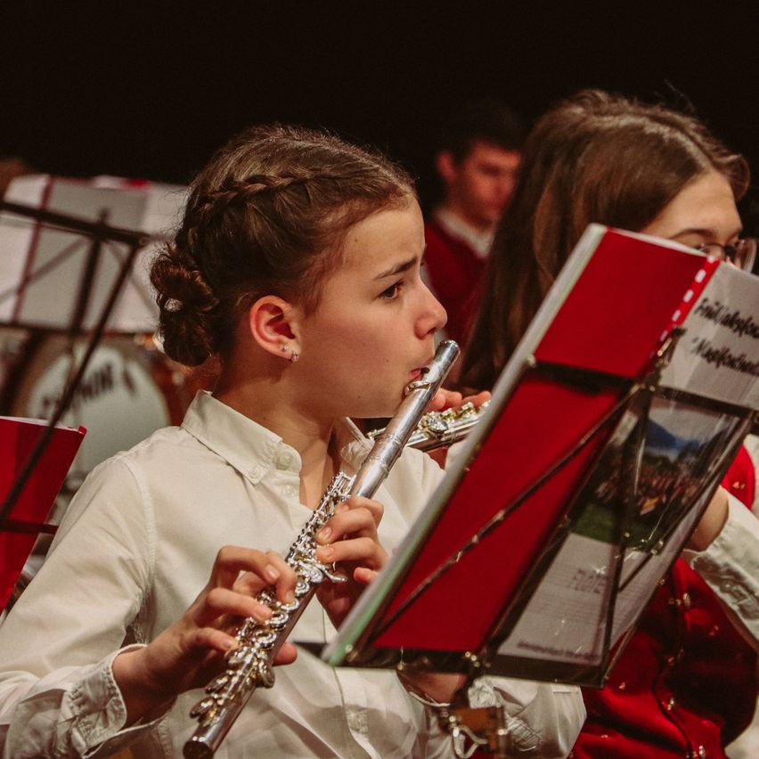 Ein Mädchen spielt Flöte und schaut in ihr Notenbuch. Ein anderes Mädchen liest hinter ihr die Musik. Sie sind in einem Schulorchester.