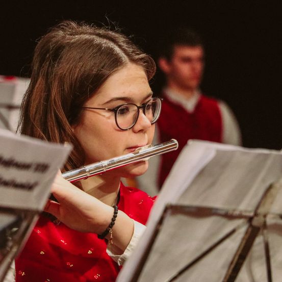 Ein Mädchen mit Brille und rotem Kleid spielt Flöte und hält Musikblätter in den Händen, mit einer verschwommenen Figur eines Mannes in einem roten West hinter ihr.