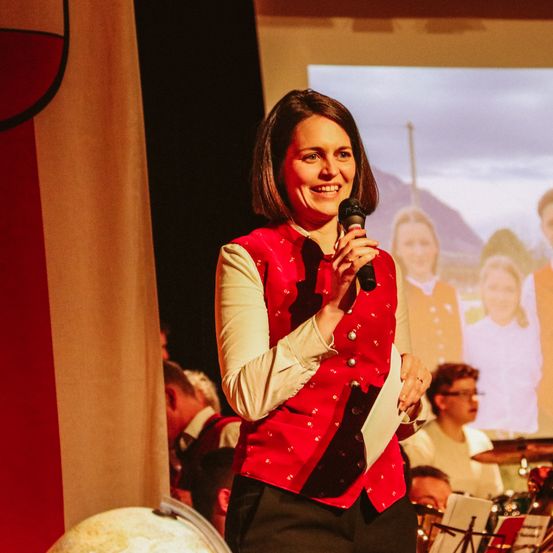 Eine Frau mit einem Mikrofon auf der Bühne bei einer Veranstaltung, sie lächelt, mit einer Flagge und einer großen Leinwand im Hintergrund, die Menschen zeigt.