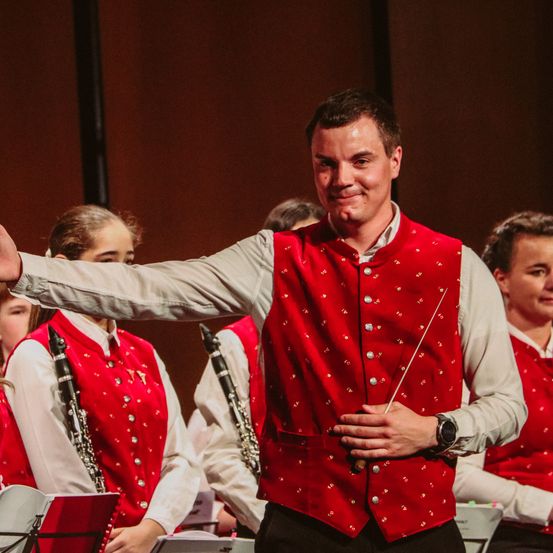 Ein Dirigent einer Band lächelt und zeigt mit seinem Taktstock auf eine Gruppe von Musikern.