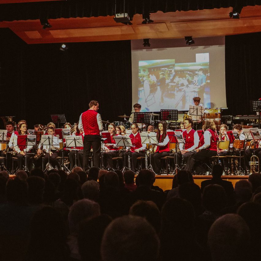 Eine Gruppe von Musikern, darunter Trompeten, Posaunen und Holzblasinstrumente, spielt auf der Bühne. Sie sind in roten Westen und schwarzen Hosen gekleidet. Im Hintergrund zeigt eine Projektionsleinwand ein Bild von Menschen in einem Raum.