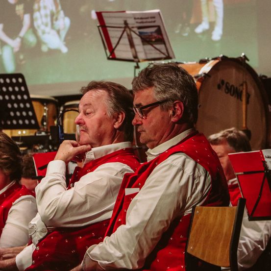 Zwei Männer in roten Westen sitzen nebeneinander im Sitzbereich einer Band. Dahinter sind ein großes Schlagzeug und ein Notenständer zu sehen.