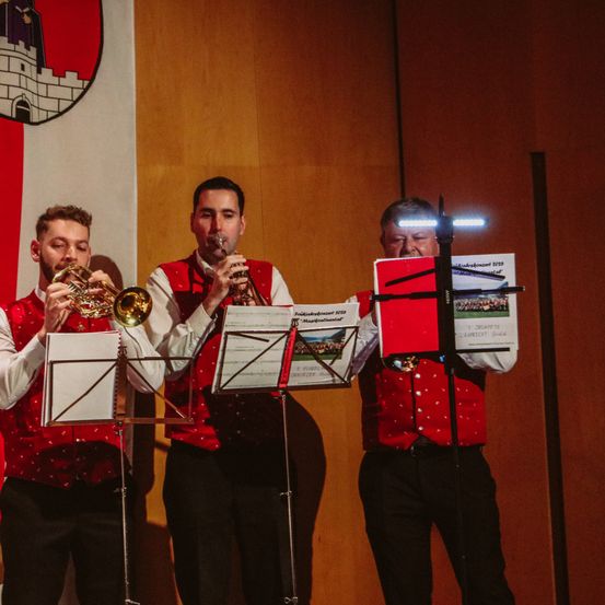 Drei Musiker spielen Trompeten auf einer Bühne, einer liest von einem Notenständer mit Lichtern darauf. Hinter ihnen hängt eine rot-weiße Fahne mit einem Logo an der Wand.