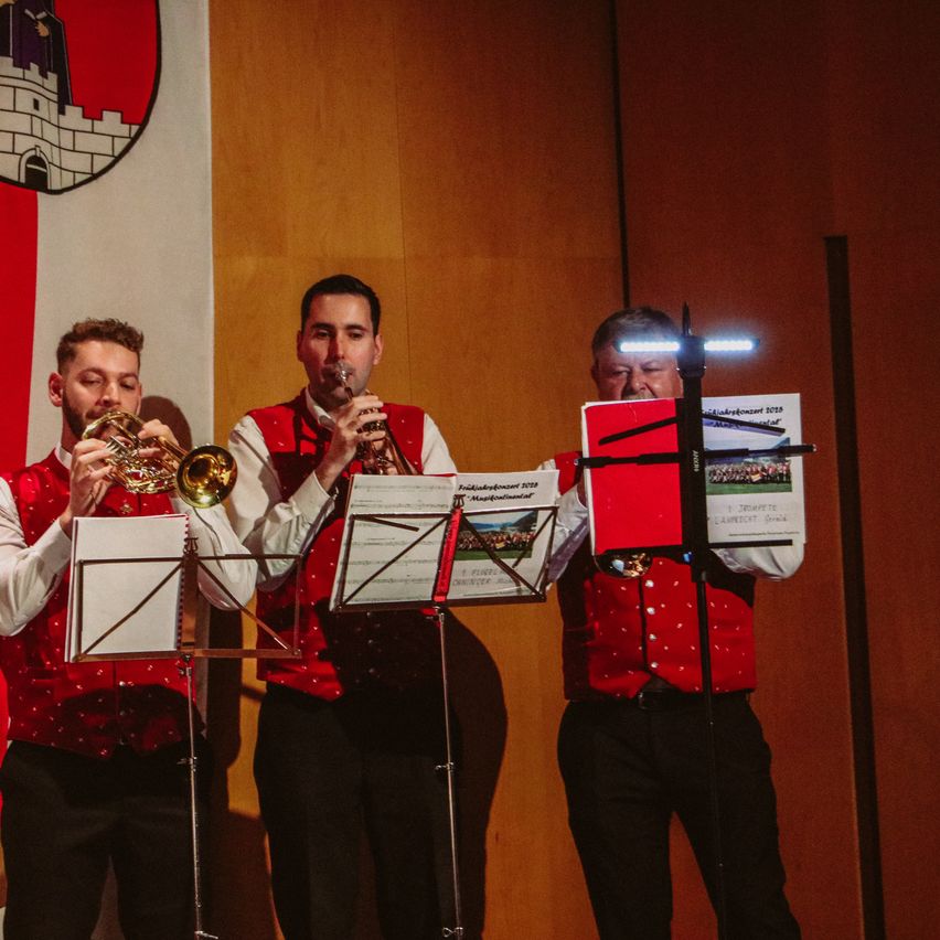 Drei Musiker spielen Trompeten auf einer Bühne, einer liest von einem Notenständer mit Lichtern darauf. Hinter ihnen hängt eine rot-weiße Fahne mit einem Logo an der Wand.