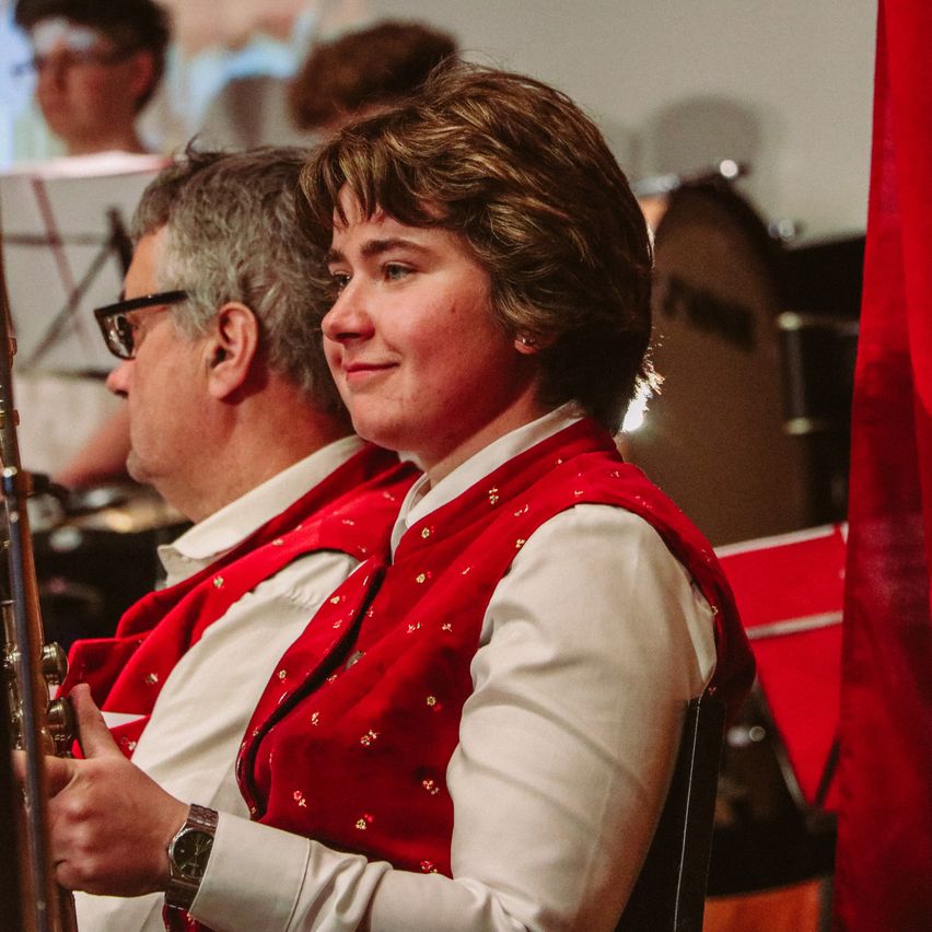 Eine Frau in einer roten Weste sitzt neben einem Mann in einer weißen Weste. Sie lächeln beide und schauen nach vorne. Hinter ihnen steht ein Schlagzeug, und eine Person sitzt mit Musikinstrumenten.