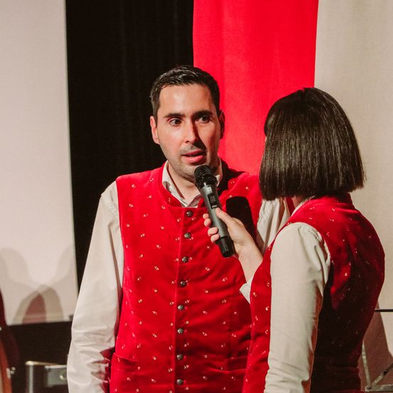 Ein Mann und eine Frau stehen vor einem roten Hintergrund, beide tragen rote Westen. Der Mann hält ein Mikrofon und schaut die Frau an, die spricht.