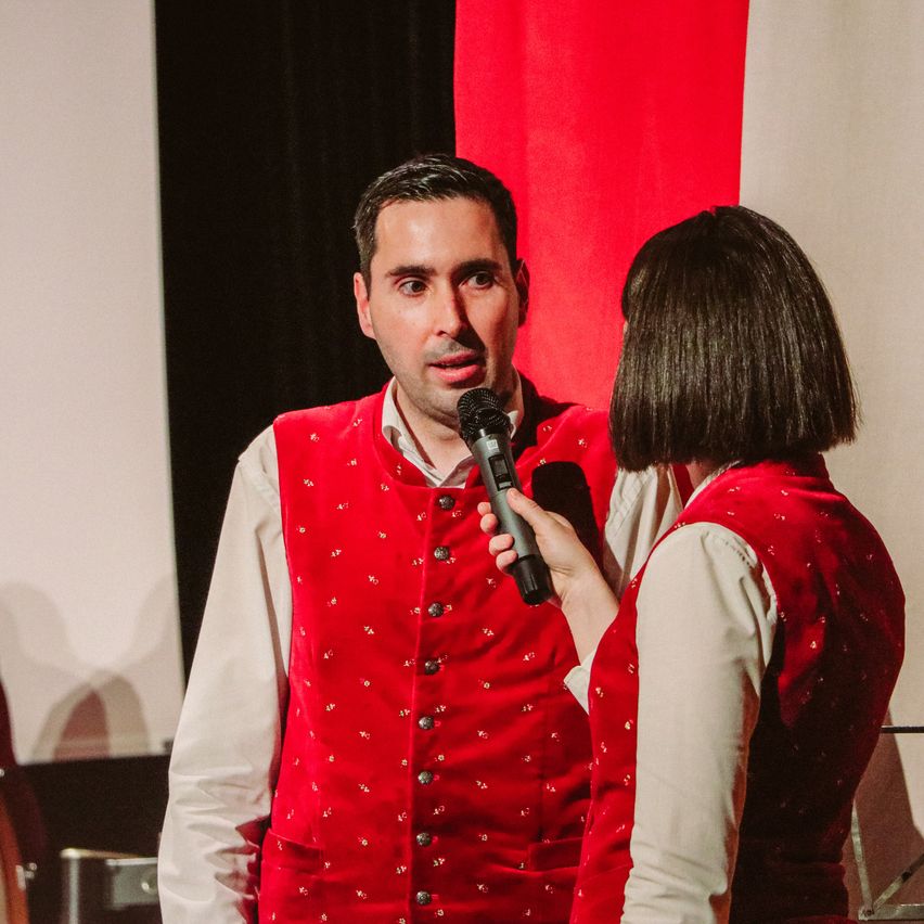 Ein Mann und eine Frau stehen vor einem roten Hintergrund, beide tragen rote Westen. Der Mann hält ein Mikrofon und schaut die Frau an, die spricht.