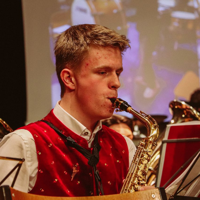 Ein junger Mann in einem roten Westchen spielt eine Saxophon und schaut darauf.