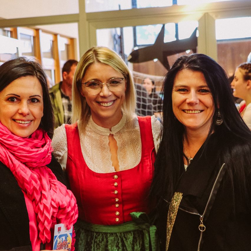 Drei lächelnde Frauen in einem Raum, eine in einem roten Dirndl und einem rosa Schal, eine andere in einem schwarzen Outfit mit einer Halskette und die dritte in einem schwarzen Mantel.