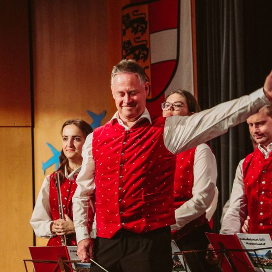 Ein Mann mit einem roten Westchen dirigiert eine Band mit einem Taktstock, vor drei Musikern mit Flöten und einer Fahne mit einem Löwen und einem roten Schild.