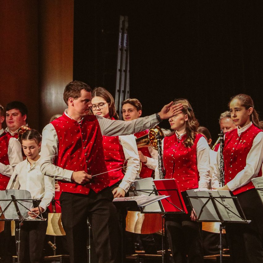 Ein Schulorchester tritt auf der Bühne auf, angeführt von einem Dirigenten, während die Musiker ihre Instrumente spielen, darunter Trompeten, Posaunen und Geigen.