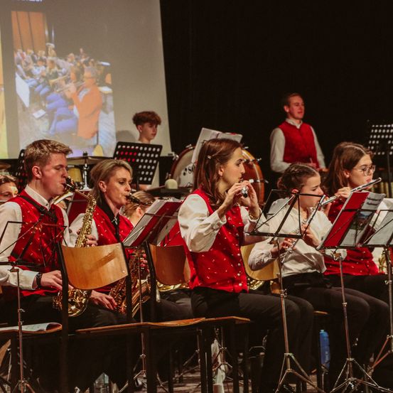 Eine Gruppe von Musikern spielt auf der Bühne, einige spielen Saxophon und andere Flöte. Die Musiker tragen rote Westen mit weißen langen Ärmeln. Dahinter zeigt eine Projektionsleinwand ein Bild einer Gruppe von Musikern, die Instrumente spielen.