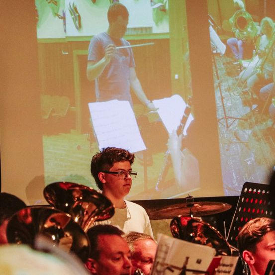 Ein junger Mann steht vor einer Leinwand, die einen Dirigenten zeigt, während mehrere andere Musiker auf dem Boden sitzen und Instrumente spielen.