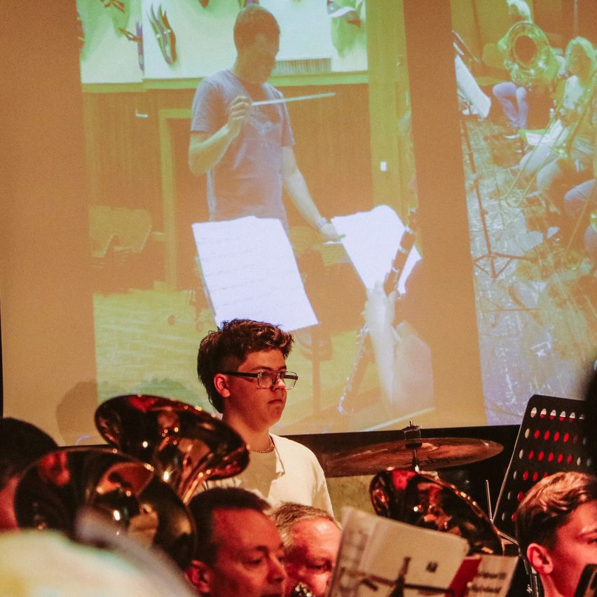 Ein junger Mann steht vor einer Leinwand, die einen Dirigenten zeigt, während mehrere andere Musiker auf dem Boden sitzen und Instrumente spielen.