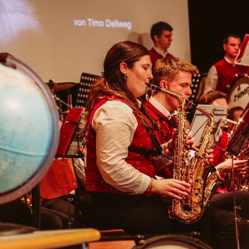 Eine Frau in rotem und weißem Outfit spielt Saxophon in einem Musikensemble. Im Vordergrund sind ein Globus und Notenständer zu sehen.