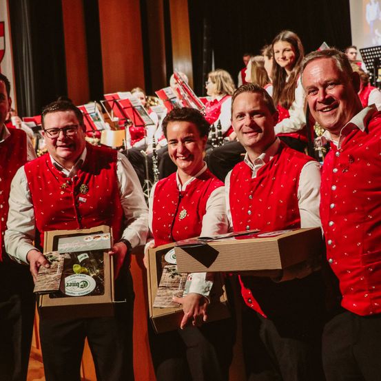 Vier erwachsene Männer, gekleidet in roten Westen, halten Boxen und lächeln in die Kamera. Dahinter stehen mehrere Menschen in roten Westen mit Musikinstrumenten.