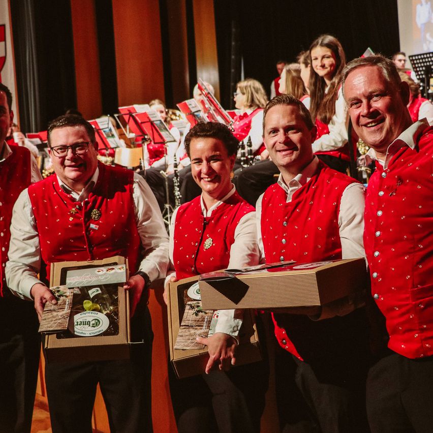 Vier erwachsene Männer, gekleidet in roten Westen, halten Boxen und lächeln in die Kamera. Dahinter stehen mehrere Menschen in roten Westen mit Musikinstrumenten.