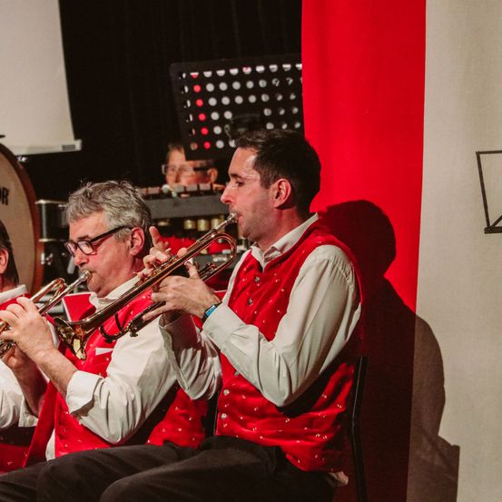 Zwei Männer in roten Westen und weißen Langarmhemden spielen Trompeten. Ein Mann sitzt auf einem Stuhl, der andere steht. Sie werden von einem Scheinwerfer auf einem roten Hintergrund beleuchtet, mit Musikinstrumenten im Hintergrund.