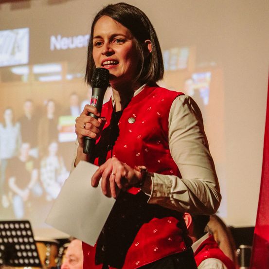 Eine Frau in einem roten Wams steht auf der Bühne und hält ein Mikrofon. Sie lächelt und scheint eine Rede zu halten. Hinter ihr zeigt eine Projektionsleinwand eine Gruppe von Menschen.