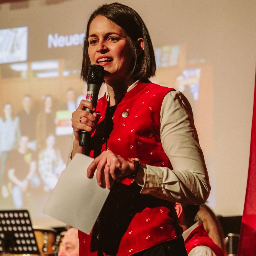 Eine Frau in einem roten Wams steht auf der Bühne und hält ein Mikrofon. Sie lächelt und scheint eine Rede zu halten. Hinter ihr zeigt eine Projektionsleinwand eine Gruppe von Menschen.