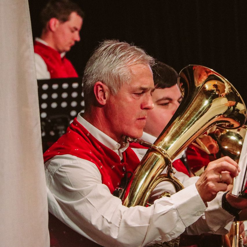 Ein Mann in einem roten Vest spielt ein Blasinstrument, möglicherweise eine Tuba, während eine andere Person im Hintergrund ebenfalls ein Blasinstrument spielt.